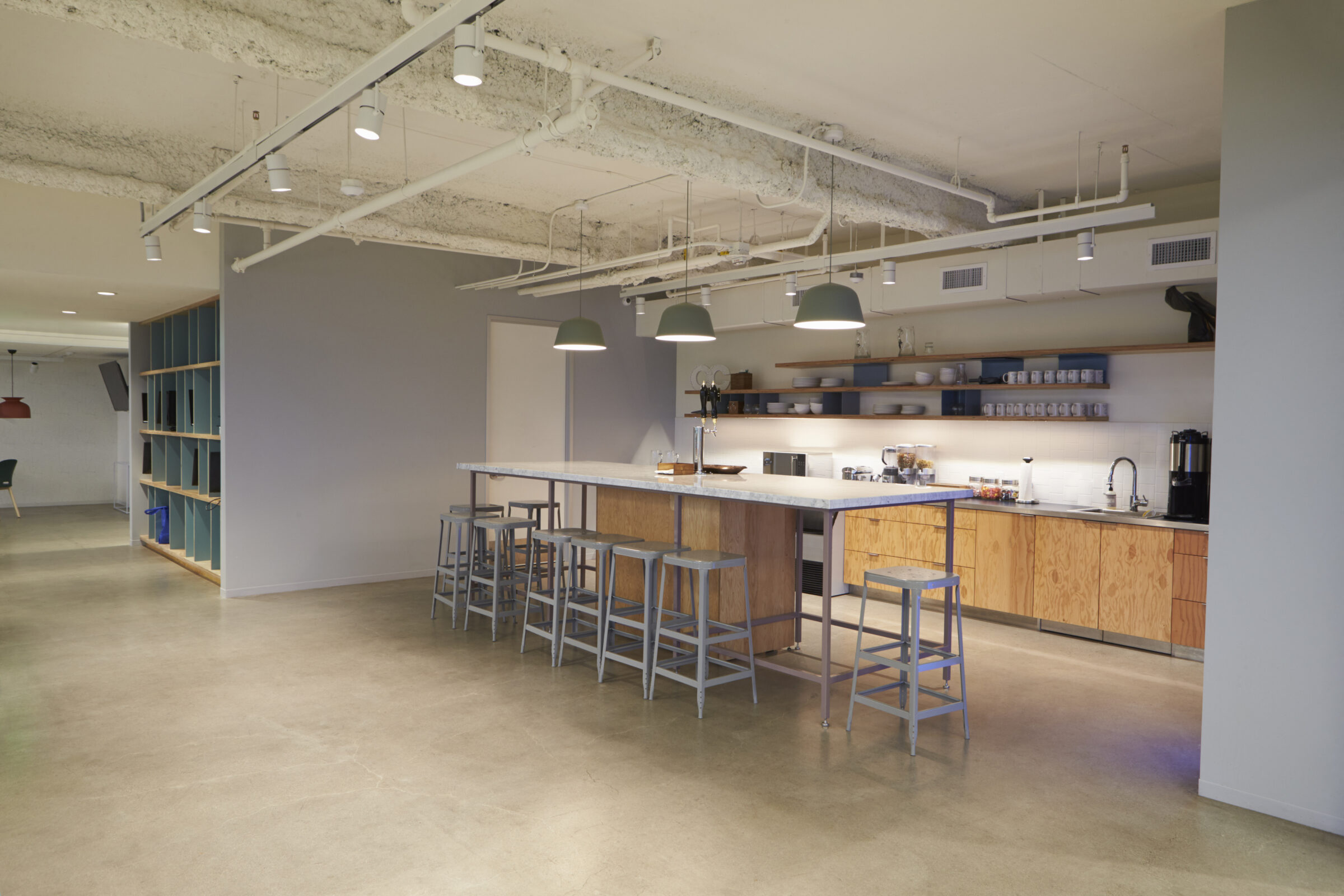 Modern office kitchen featuring a long island with bar stools, industrial lighting, shelves with mugs, and exposed ceiling with pipes.