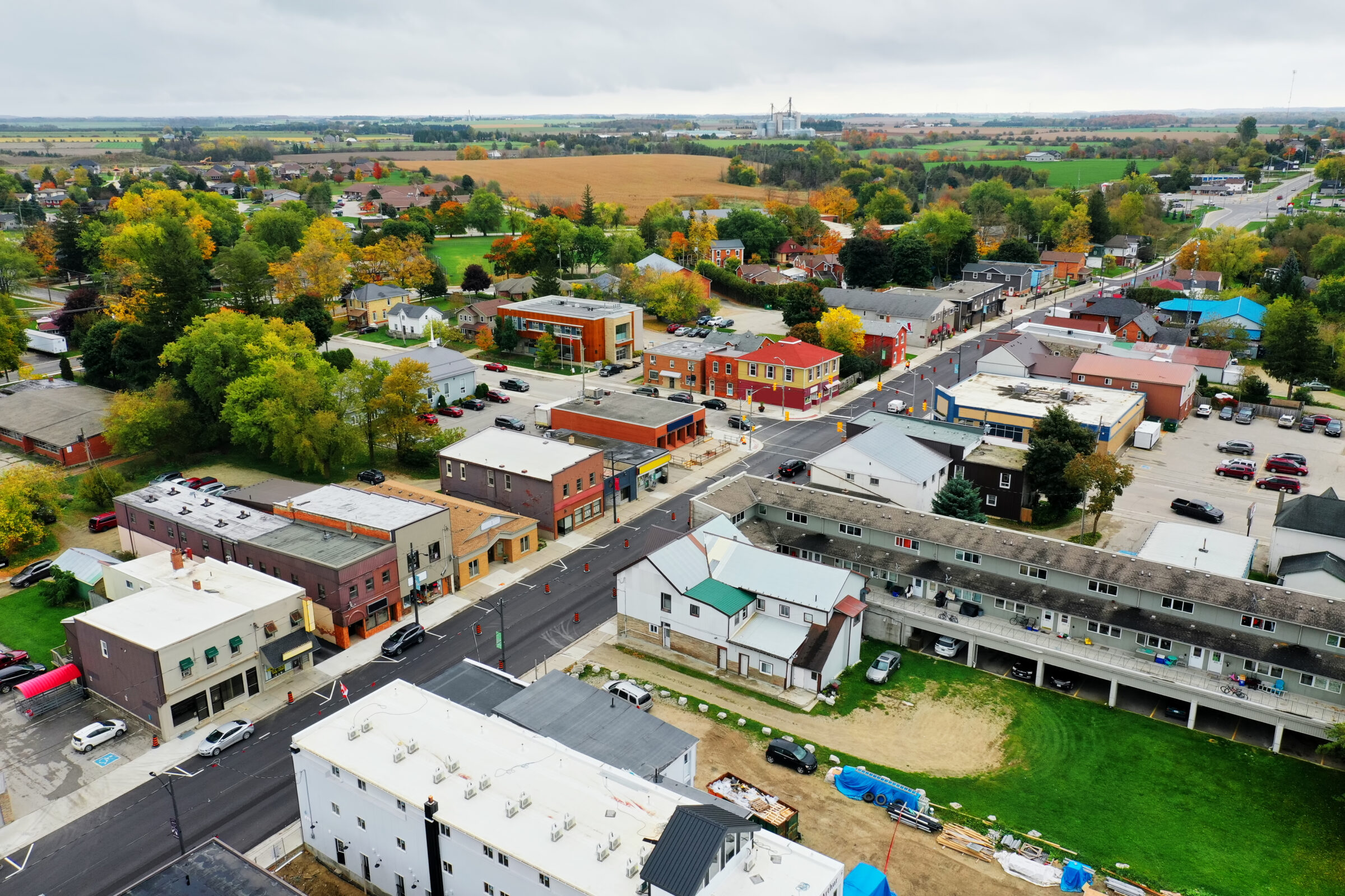 Aerial view of a small town with colorful buildings, surrounded by trees and fields, featuring a central road and residential areas.