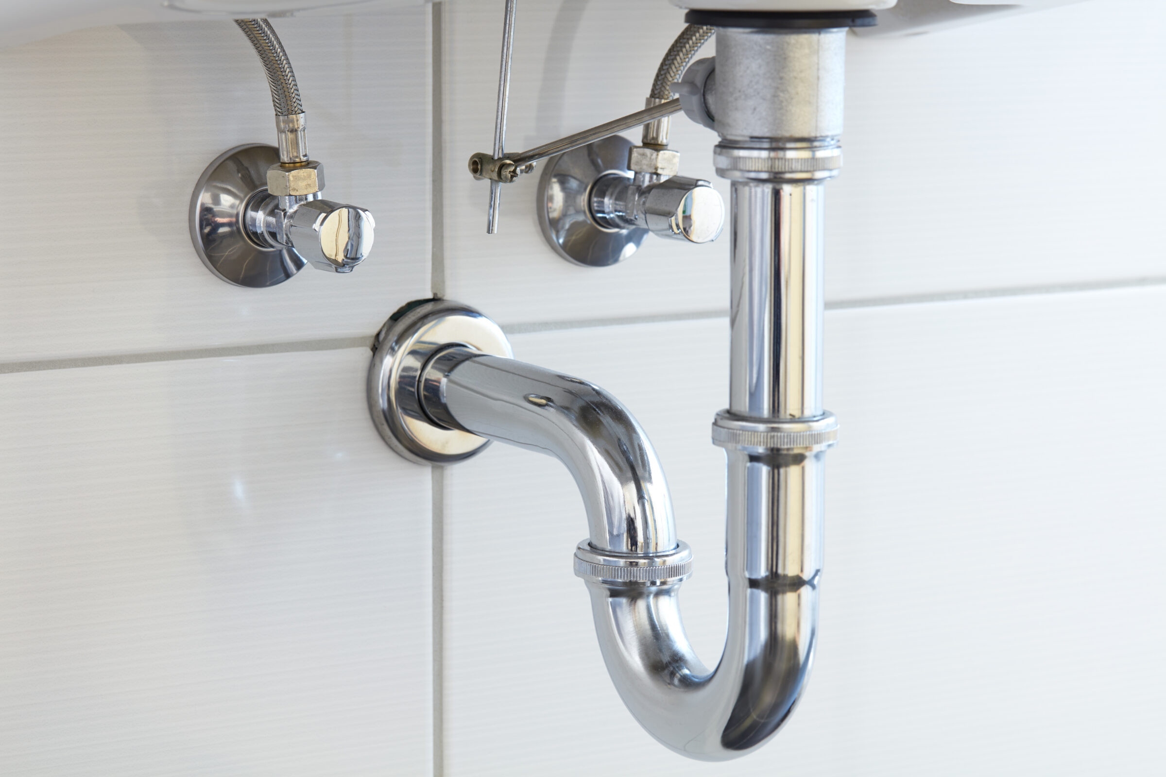 Chrome plumbing pipes and fixtures beneath a white tiled sink, showcasing a clean and modern under-sink setup in a bathroom or kitchen.