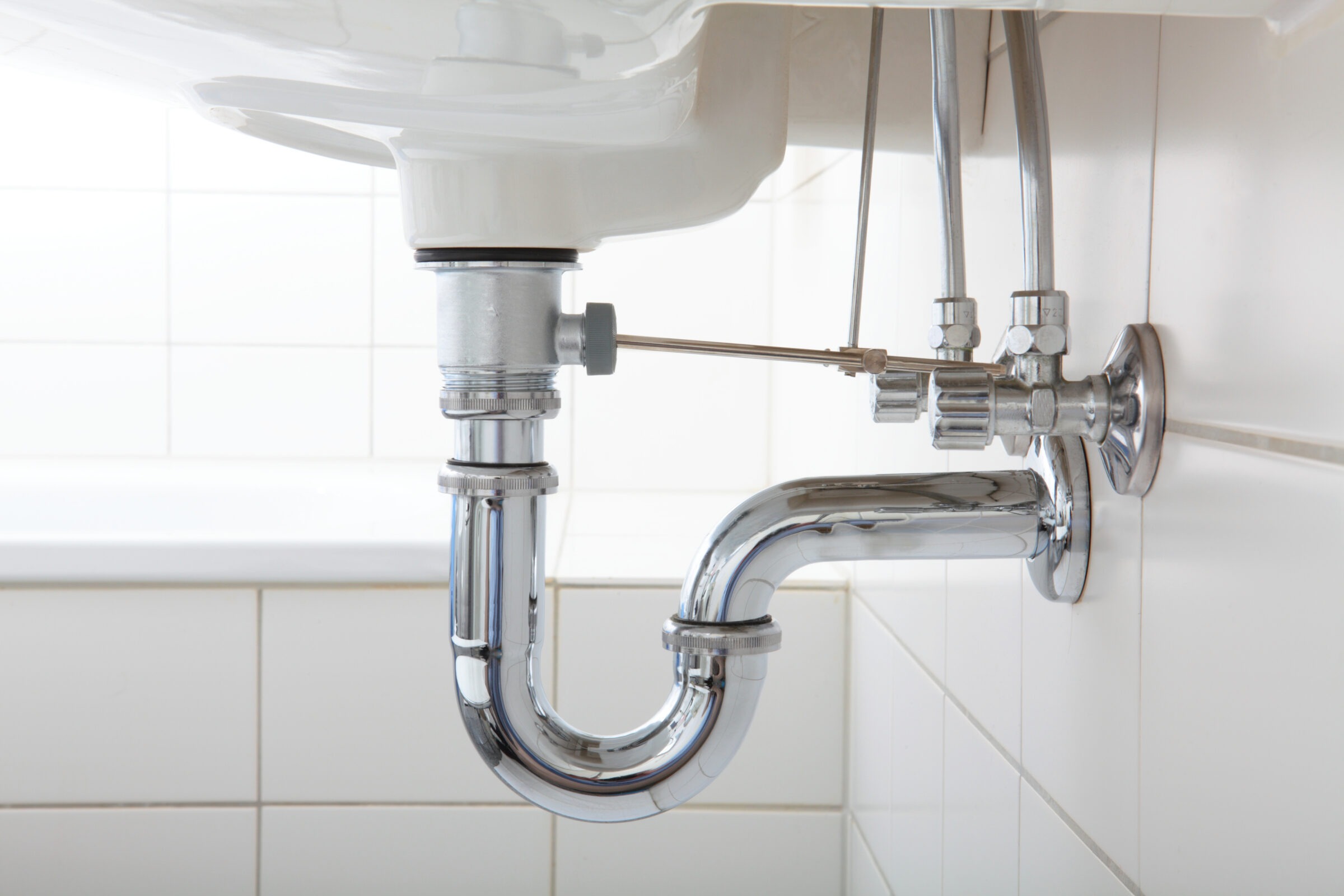 A shiny chrome sink plumbing system below a white basin on tiled wall, featuring U-bend pipes and valve connections. Clean and functional setup.